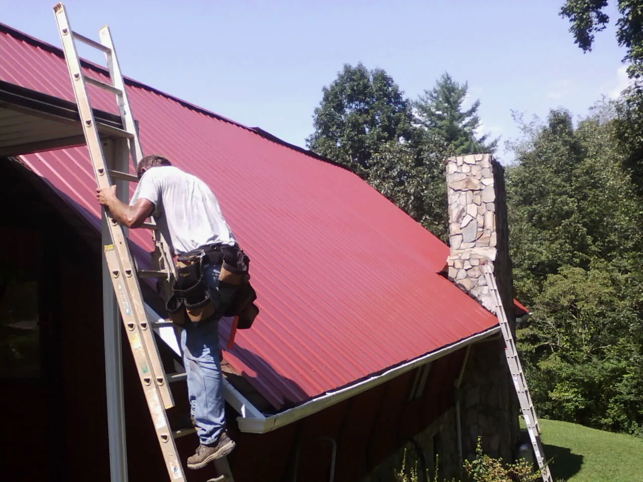 Roofer on a ladder beside a red metal roof and stone chimney — Soffit & Fascia Repair by Roofing Brothers in Cedar Vale